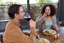 Felice coppia diversificata in soggiorno seduto a tavola cenare insieme e bere vino. trascorrendo del tempo a casa in un appartamento moderno. — Foto stock