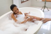 Змішана расова жінка у ванній має ванну і голиться ногами. домашній спосіб життя, насолоджуючись доглядом за дозвіллям вдома . — стокове фото