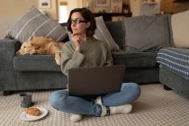 Mujer caucásica en la sala de estar con su perro mascota, sentado en el suelo, trabajando con el ordenador portátil. estilo de vida doméstico, disfrutando del tiempo libre en casa. - foto de stock