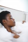 Femme afro-américaine souriante dans la salle de bain, relaxante dans la baignoire avec les yeux fermés. mode de vie domestique, profiter de loisirs d'auto-soins à la maison. — Photo de stock