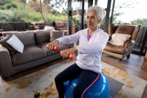 Heureuse femme caucasienne âgée dans le salon faisant de l'exercice, assise sur des haltères de levage de boules suisses. mode de vie à la retraite, passer du temps seul à la maison. — Photo de stock