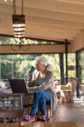Entspannte ältere kaukasische Frau in der Küche mit Laptop und Kaffee trinken. Lebensstil im Ruhestand, Zeit allein zu Hause verbringen. — Stockfoto