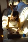 Змішана жінка, що лежить і використовує ноутбук у сонячній вітальні. здоровий спосіб життя, насолоджуючись дозвіллям вдома . — стокове фото