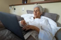 Mulher caucasiana sênior no quarto, sentada na cama e usando laptop. estilo de vida aposentadoria, passar o tempo sozinho em casa. — Fotografia de Stock