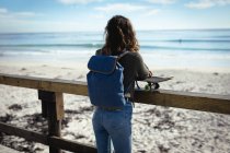 Mujer de raza mixta sosteniendo monopatín en un día soleado junto al mar. estilo de vida saludable, disfrutar del tiempo libre al aire libre. - foto de stock