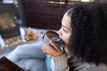 Feliz mujer de raza mixta en la sala de estar sentado junto a la chimenea sosteniendo taza y beber café. pasar tiempo libre en casa. . - foto de stock