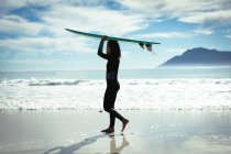 Mujer de raza mixta sosteniendo tabla de surf en un día soleado en la playa. estilo de vida saludable, disfrutar del tiempo libre al aire libre. - foto de stock