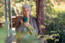Relajante mujer mayor caucásica en el balcón de pie y beber café. estilo de vida de jubilación, pasar tiempo solo en casa. - foto de stock