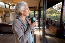 Femme caucasienne âgée dans la cuisine regardant la fenêtre et buvant du café. mode de vie à la retraite, passer du temps seul à la maison. — Photo de stock
