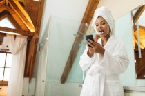 Mixed Race Frau im Badezimmer mit Smartphone und Zähneputzen. häuslicher Lebensstil, selbstgepflegte Freizeit zu Hause genießen. — Stockfoto
