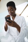 Mujer afroamericana en baño cepillándose los dientes y usando smartphone. estilo de vida doméstico, disfrutando del tiempo libre de autocuidado en casa. - foto de stock