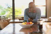 Homem americano africano sênior localizado na cozinha moderna e usando smartphone. estilo de vida aposentadoria, passar o tempo sozinho em casa. — Fotografia de Stock