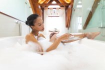 Mujer de raza mixta en el baño tomando un baño y afeitándose las piernas. estilo de vida doméstico, disfrutando del tiempo libre de autocuidado en casa. - foto de stock
