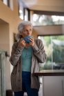 Mujer caucásica mayor en la cocina de pie y tomando café. estilo de vida de jubilación, pasar tiempo solo en casa. - foto de stock