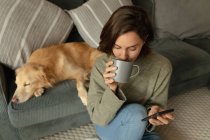 Mujer caucásica en la sala de estar con su perro mascota, el uso de teléfonos inteligentes y beber café. estilo de vida doméstico, disfrutando del tiempo libre en casa. - foto de stock