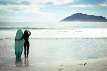 Mujer de raza mixta sosteniendo tabla de surf en el mar en el día soleado. estilo de vida saludable, disfrutar del tiempo libre al aire libre. - foto de stock