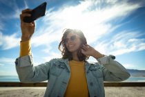 Mujer de raza mixta tomando selfie con smartphone en un día soleado junto al mar. estilo de vida saludable, disfrutar del tiempo libre al aire libre. - foto de stock