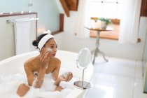 Смешанная расовая женщина в ванной комнате, принимающая ванну и надевающая маску красоты. домашний образ жизни, наслаждаясь отдыхом на дому. — стоковое фото
