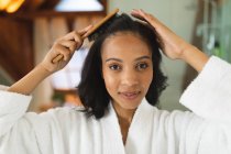 Portrait de femme souriante métissée dans la salle de bain brossant les cheveux en regardant la caméra. mode de vie domestique, profiter de loisirs d'auto-soins à la maison. — Photo de stock