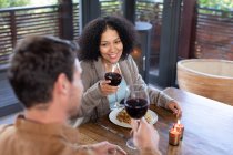 Feliz casal diversificado na sala de estar sentado à mesa jantando juntos e bebendo vinho. passar o tempo fora em casa no apartamento moderno. — Fotografia de Stock