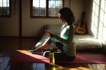 Femme de race mixte pratiquant le yoga, en utilisant un smartphone dans le salon ensoleillé. mode de vie sain, profiter de loisirs à la maison. — Photo de stock