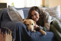 Портрет улыбающейся белой женщины в гостиной, сидящей на диване и обнимающей свою собаку. домашний образ жизни, наслаждаясь отдыхом дома. — стоковое фото