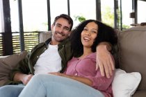 Retrato de feliz pareja diversa sentada en el sofá en la sala de estar abrazando y sonriendo. pasar tiempo libre en casa en apartamento moderno. - foto de stock