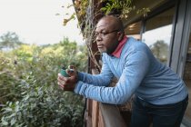Homme afro-américain senior réfléchi sur un balcon ensoleillé versant une tasse de café. mode de vie à la retraite, passer du temps seul à la maison. — Photo de stock