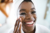 Портрет улыбающейся африканской американки в ванной комнате, наносящей крем для лица для ухода за кожей. домашний образ жизни, наслаждаясь отдыхом на дому. — стоковое фото
