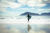 Змішана расова жінка тримає дошку для серфінгу в морі в сонячний день. здоровий спосіб життя, насолоджуючись дозвіллям на відкритому повітрі . — стокове фото