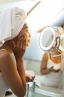 Mixed race woman in bathroom applying face cream for skin care, looking in mirror. domestic lifestyle, enjoying self care leisure time at home. — Stock Photo