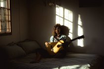 Gemischte Rasse Frau spielt Gitarre im sonnigen Schlafzimmer. Gesunder Lebensstil, Freizeit zu Hause genießen. — Stockfoto