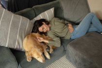 Mulher branca sorridente na sala de estar, deitada no sofá com seu cachorro de estimação. estilo de vida doméstico, desfrutando de tempo de lazer em casa. — Fotografia de Stock