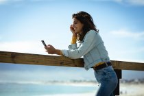 Змішана расова жінка використовує смартфон у сонячний день на узбережжі. здоровий спосіб життя, насолоджуючись дозвіллям на відкритому повітрі . — стокове фото