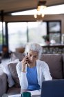 Старша біла жінка у вітальні сидить на дивані, використовуючи смартфон і ноутбук. пенсійний спосіб життя, проводити час наодинці вдома . — стокове фото