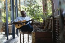 Détente homme afro-américain senior sur un balcon ensoleillé jouant de la guitare. mode de vie à la retraite, passer du temps seul à la maison. — Photo de stock