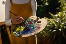 Caucasian woman in sunny garden, mixing paint on palette. domestic lifestyle, enjoying leisure time at home. — Stock Photo