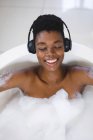 Lächelnde Afroamerikanerin entspannt sich im Bad mit geschlossenen Augen und hört Musik über Kopfhörer. — Stockfoto