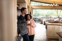 Felice coppia diversificata in cucina abbracciando bere caffè e sorridente. trascorrendo del tempo a casa in un appartamento moderno. — Foto stock