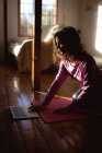 Mujer de raza mixta practicando yoga, utilizando portátil en la sala de estar soleada. estilo de vida saludable, disfrutando del tiempo libre en casa. - foto de stock