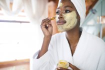 Sorrindo mulher afro-americana no banheiro aplicando máscara facial beleza. estilo de vida doméstico, desfrutando de tempo de lazer auto-cuidado em casa. — Fotografia de Stock