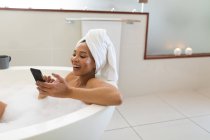 Laughing mixed race woman in bathroom having a bath and using smartphone. domestic lifestyle, enjoying self care leisure time at home. — Stock Photo
