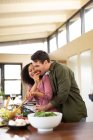 Feliz casal diversificado na cozinha preparando comida juntos comendo e sorrindo. passar o tempo fora em casa no apartamento moderno. — Fotografia de Stock