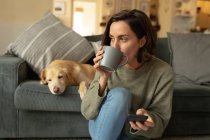 Кавказька жінка у вітальні зі своїм домашнім собакою, користується смартфоном і п'є каву. Домашнє життя, дозвілля вдома. — стокове фото