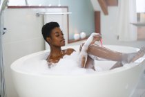 Sonriente mujer afroamericana tomando un baño de espuma y afeitándose las piernas. estilo de vida doméstico, disfrutando del tiempo libre de autocuidado en casa. - foto de stock