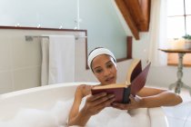 Счастливая женщина смешанной расы в ванной комнате, расслабляясь в ванной книге для чтения. домашний образ жизни, наслаждаясь отдыхом на дому. — стоковое фото