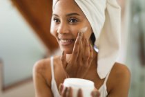 Mujer de raza mixta en baño aplicando crema facial para el cuidado de la piel, sonriendo. estilo de vida doméstico, disfrutando del tiempo libre de autocuidado en casa. - foto de stock