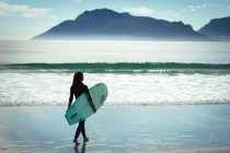Смешанные гонки женщина ходит и держит доску для серфинга на пляже в солнечный день. здоровый образ жизни, наслаждаясь отдыхом на открытом воздухе. — стоковое фото