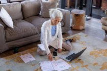 Eine ältere kaukasische Frau im Wohnzimmer sitzt auf dem Boden und arbeitet am Laptop. Lebensstil im Ruhestand, Zeit allein zu Hause verbringen. — Stockfoto
