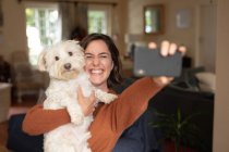 Lächelnde Kaukasierin im Wohnzimmer, die ihren Hund umarmt und ein Selfie macht. häuslicher Lebensstil, Freizeit zu Hause genießen. — Stockfoto